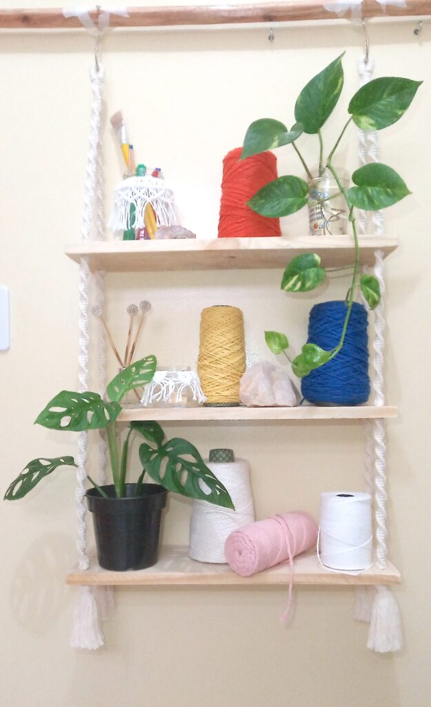 Bookcase with 3 shelves, suspended with handmade sisal rope. Rolls of colored string on the bookcase Foliage of Brazilian flora vase with flowers, handicraft material, stones and brushes.
