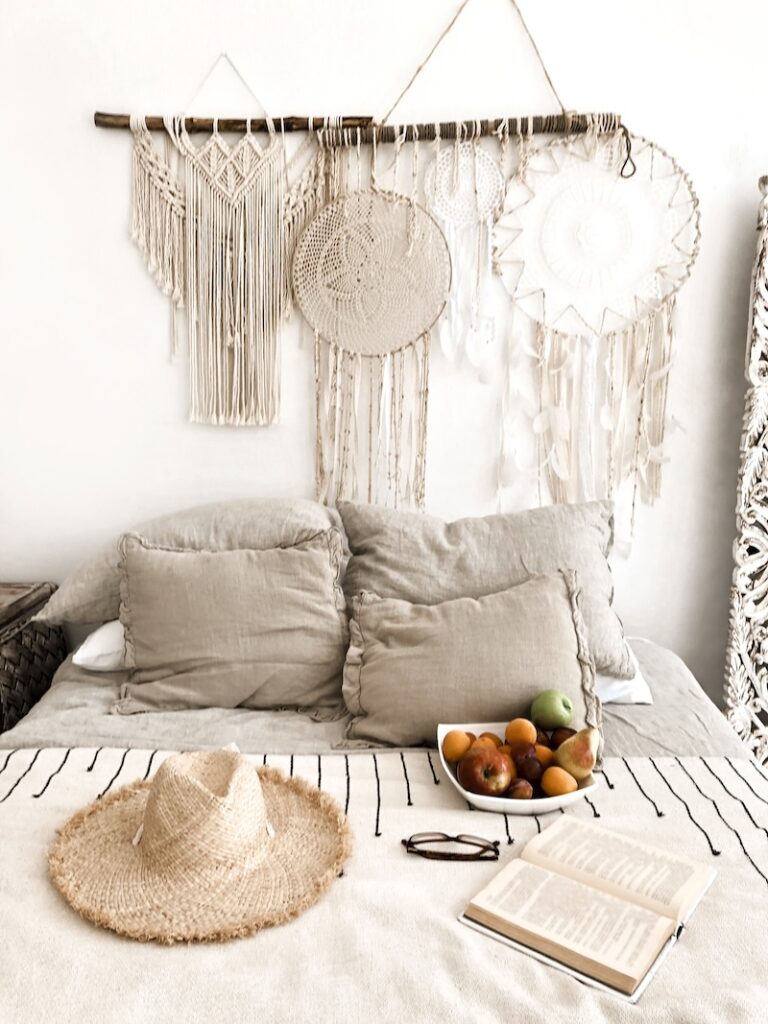 Modern scandi boho style bedroom interior with decorative pillows, wicker hat, fruits and open book on bed, macrame decorations and dreamcatcher hanging on wall panel. Light warm home atmosphere