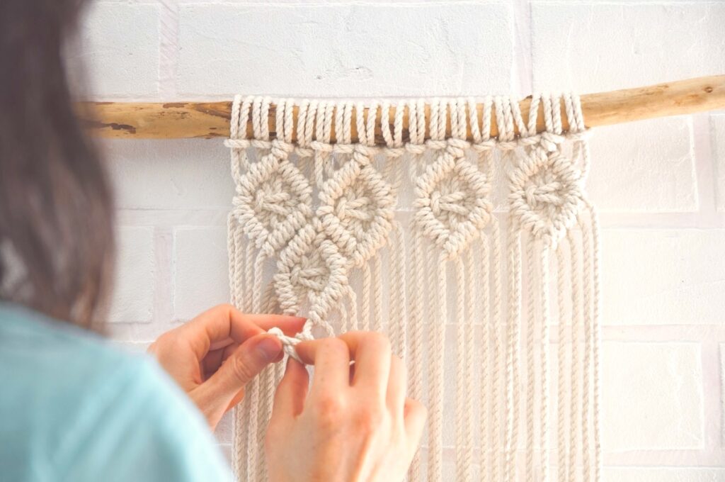 Woman making Boho wall mural made of natural color cotton threads using macrame technique.